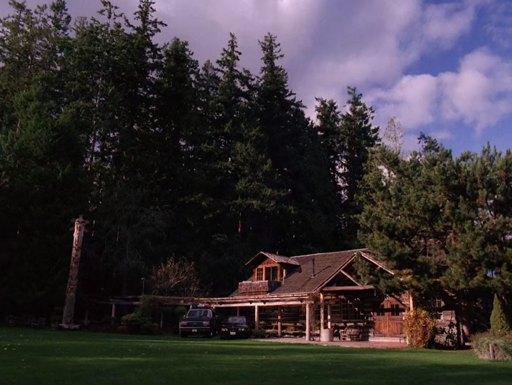 Daytime image of a wooden lodge under tall fir trees with two cars parked outside