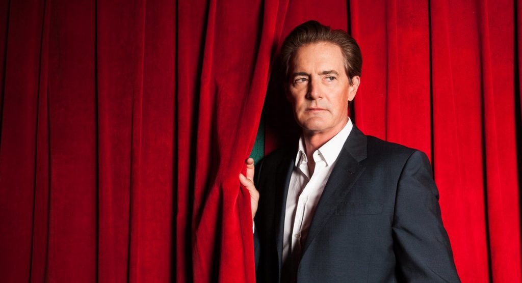 Kyle MacLachlan standing by red curtains