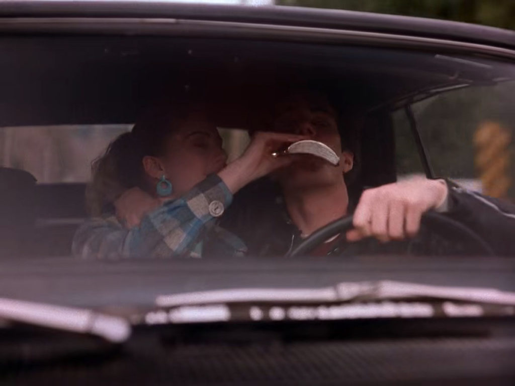 Shelly pouring a drink from a flask down Bobby's throat while he drives