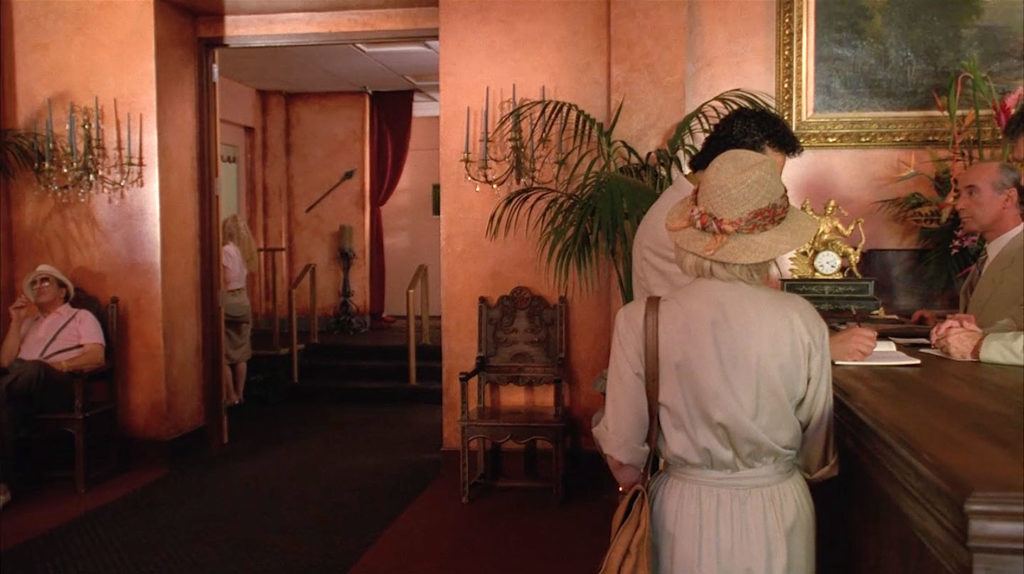 A couple is at the front desk of the Palm Deluxe Hotel while the camera looks down a hallway. A gentleman in a hat is sitting in a chair near the hall