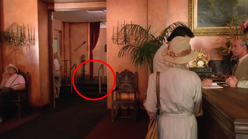 A couple is at the front desk of the Palm Deluxe Hotel while the camera looks down a hallway. A gentleman in a hat is sitting in a chair near the hall. A red circle indicates the spot where David Bowie's red shoes are seen.