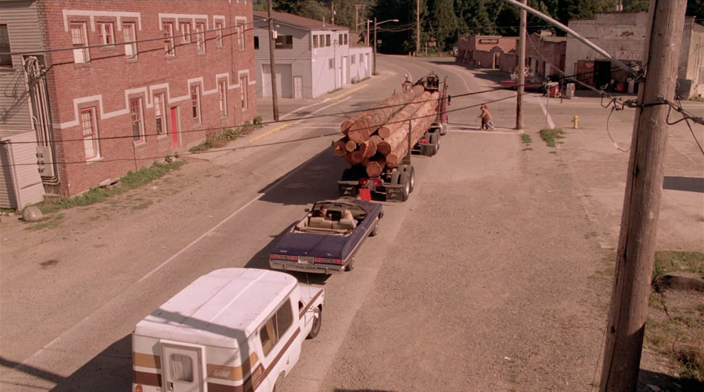 Aerial shot of cars lined up on the road behind a logging truck filled with logs