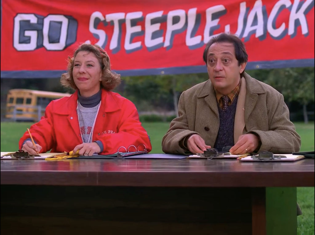 P.E. Teach and Assistant Principal Greege sit at a table with a "Go Steeplejacks" banner behind them