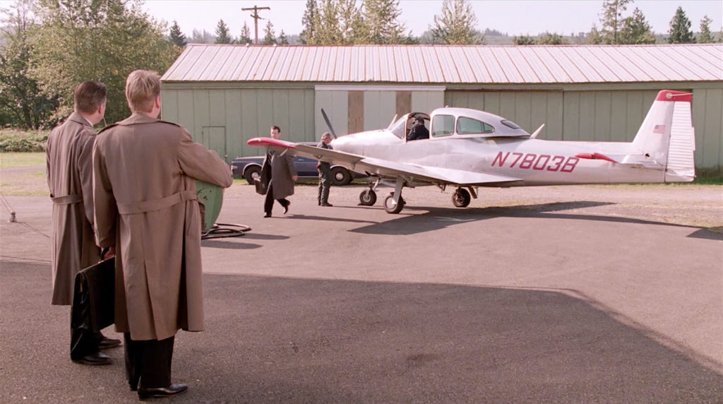 Two people waiting for someone holding a suitcase walking away from an airplane