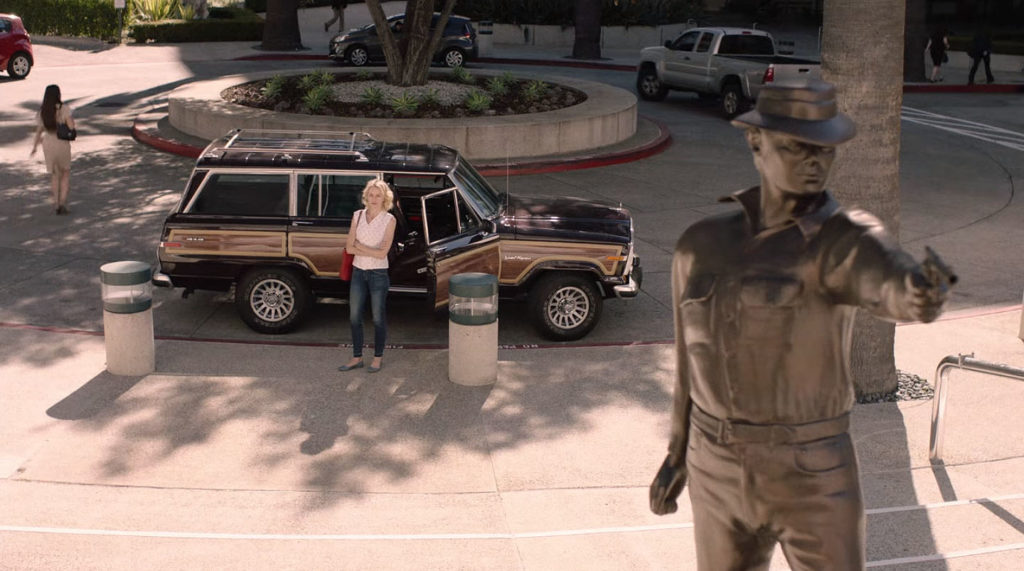 Janey-E waits by car in roundabout with statue of man holding a pistol in foreground.