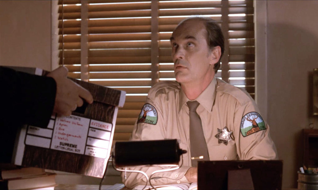 Sheriff Cable sitting behind a desk which Agent Desmond holding the evidence box