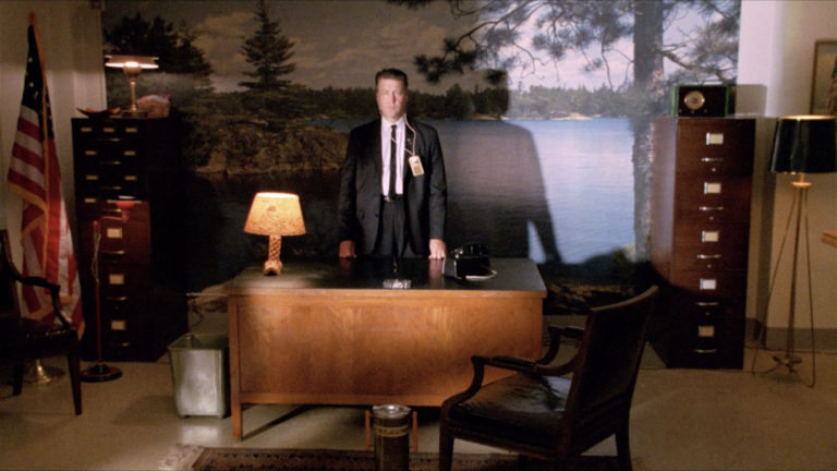 Gordon Cole standing at his desk in front of a mural