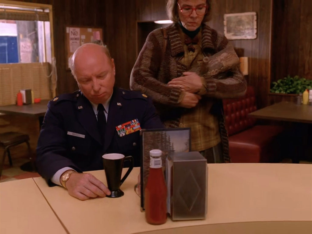Major Briggs at the Double R Diner counter while the Log Lady approaches wearing a sweater and carrying a log.