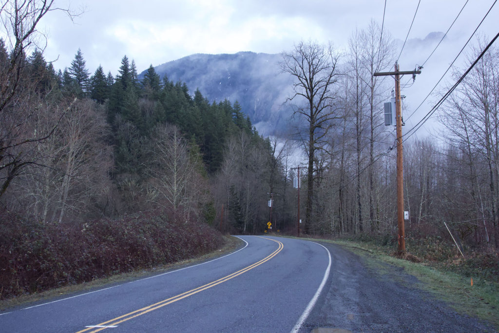 Welcome to Twin Peaks sign spot along Reinig Road in Snoqualmie, Washington. Fog covered mountains are in the distance towering above a tree-lined road.