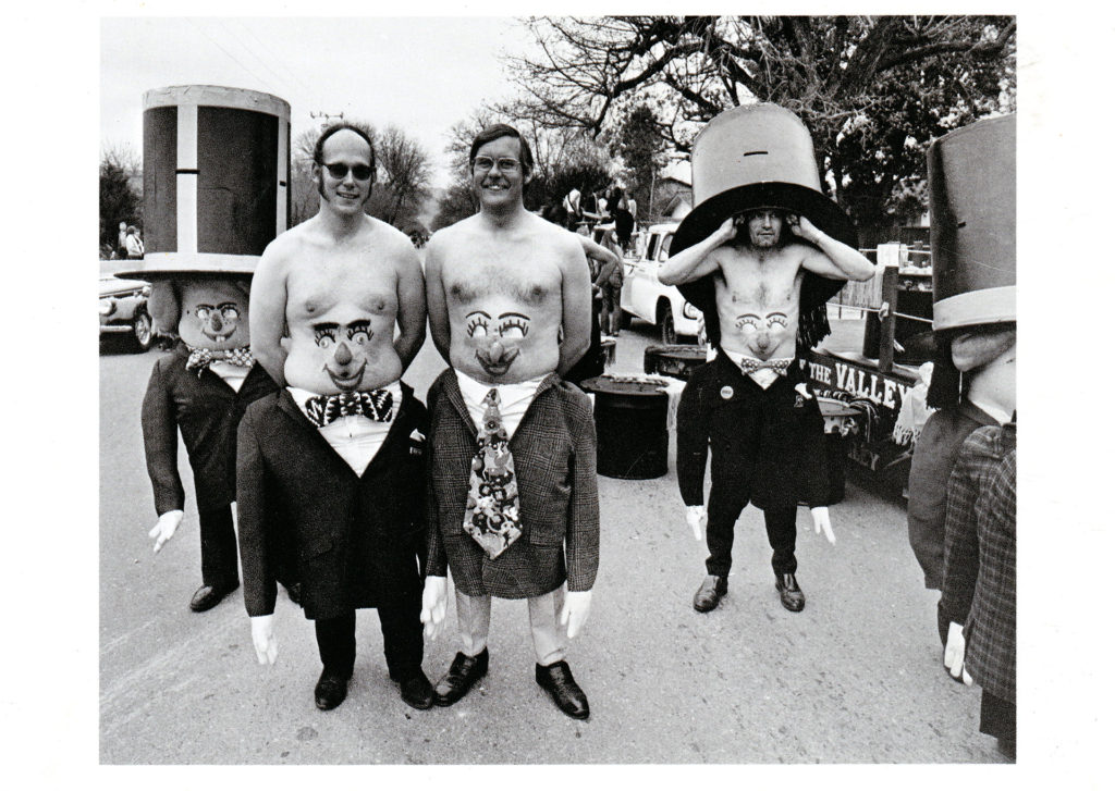Bill Owens Postcard of men standing in a parade with faces painted on their bellies 