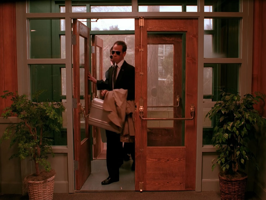 Albert Rosenfield and team arrive in Twin Peaks through the main entrance of the Twin Peaks Sheriff's Department holding tan jackets and a silver briefcase.