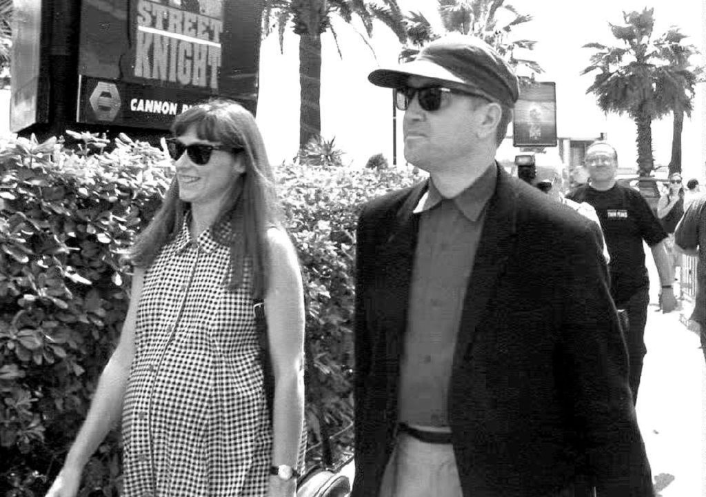 Black and white photo of David Lynch and Mary Sweeney walking on the street in Cannes. Mary is wearing a dress and David has a black suit jacket, hat and button down shirt. Both are wearing sunglasses.