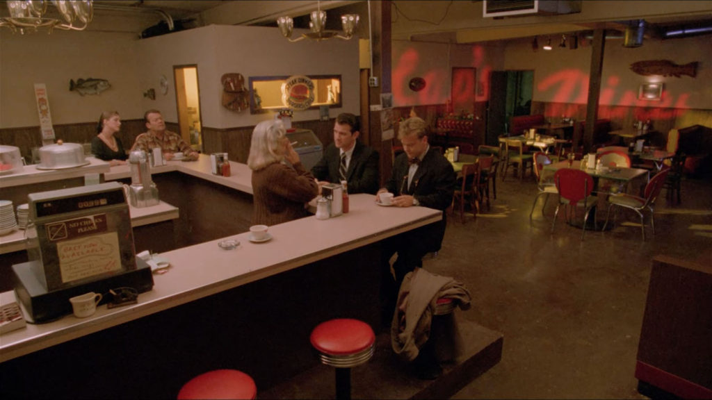 Wide shot of in the interior of Hap's Diner in Twin Peaks - Fire Walk With Me. The counter bar is seen in the foreground as FBI agents take a seat. An old man and a women sit along one side.
