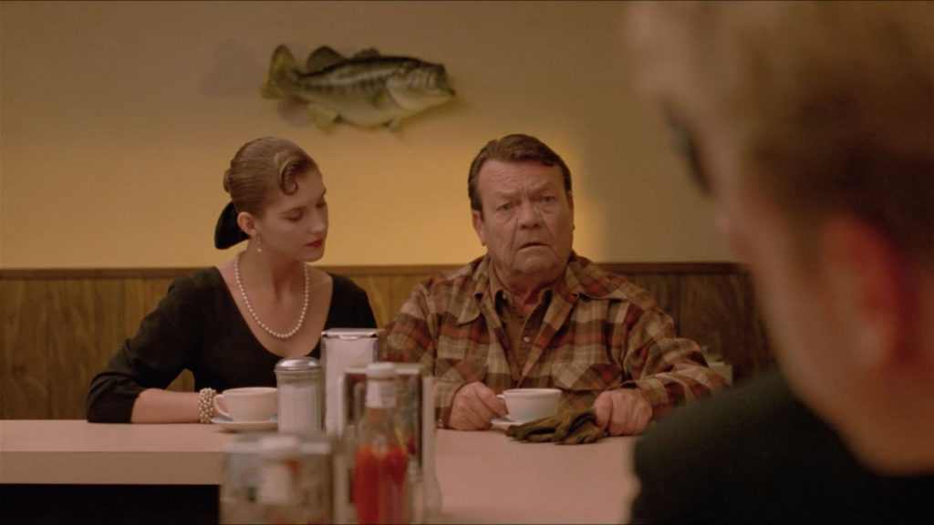 A French Girl and and Old Guy sit at the end of the counter inside Hap's while two agents listen to him speak.