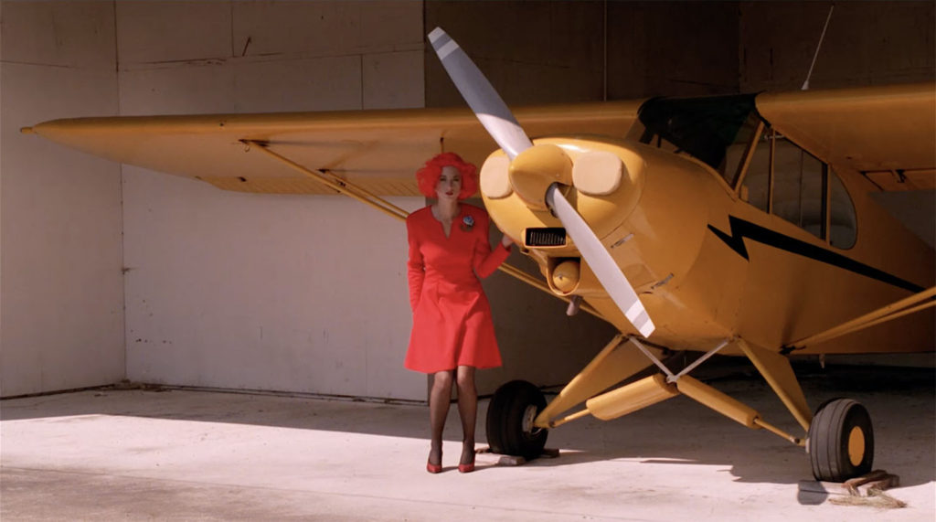 Lil the dancer in red wig and dress standing next to a yellow airplane