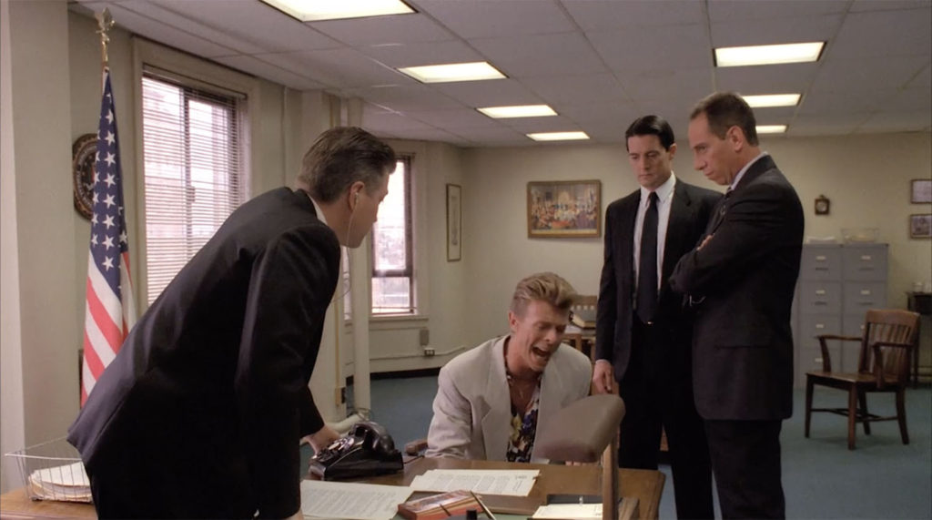 Special Agent Phillip Jeffries (David Bowie) sitting surrounded by Agent Cole (David Lynch), Agent Cooper (Kyle MacLachlan) and Agent Rosenfield (Miguel Ferrer) in the FBI Office set