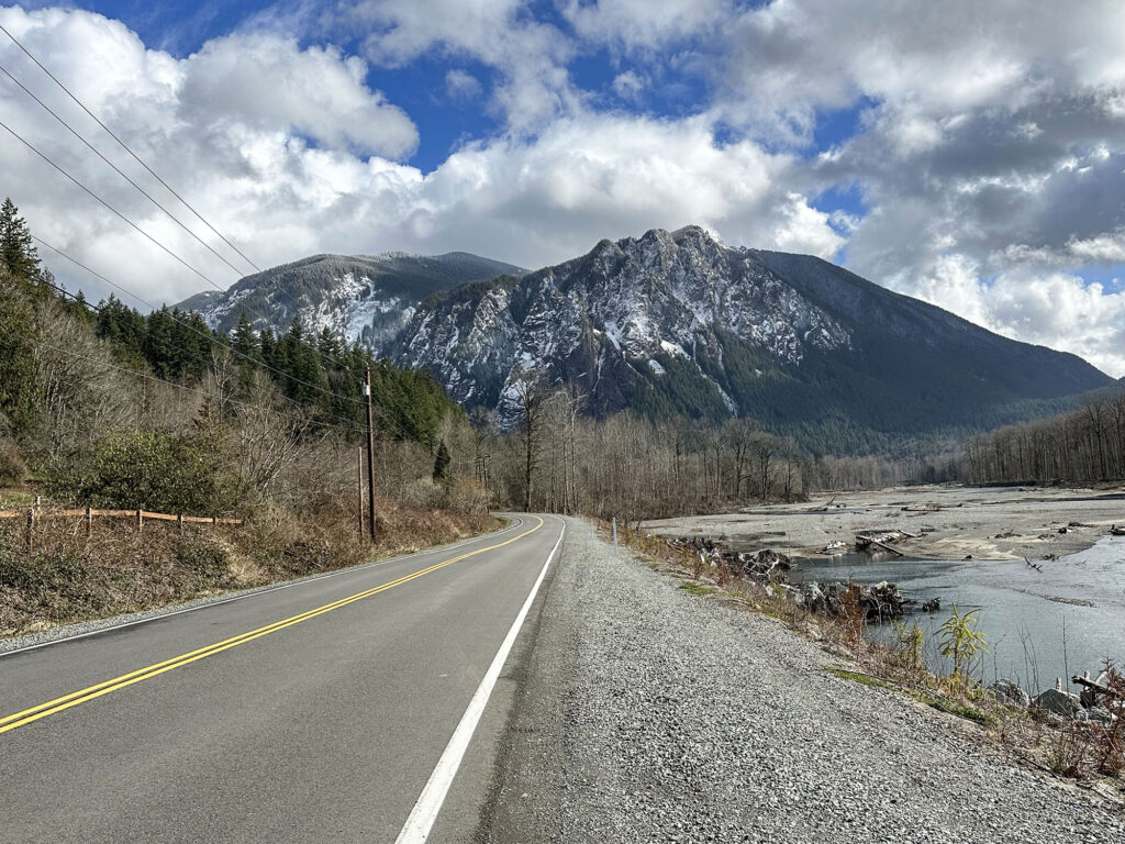 Snow capped Mount Si under cloud-covered blue skies with Reinig Road leading toward the mountain in Snoqualmie, Washington