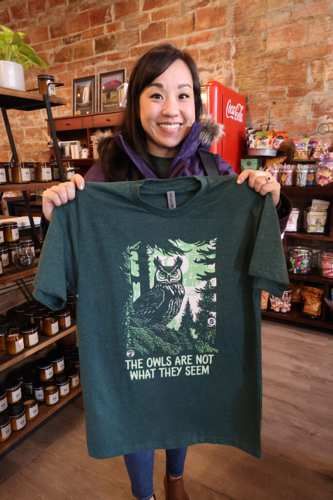 Katie holding a green t-shirt with an owl and the words "The Owls are Not What They Seem"