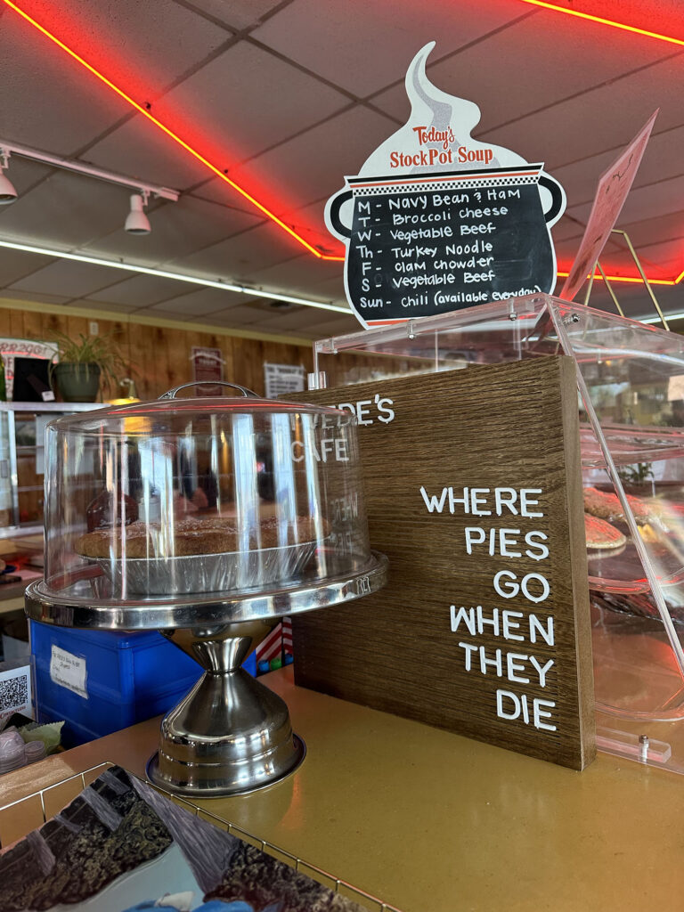 Inside of Twede's Cafe with a pie container, a wooden sign with words "Twede's Cafe - Where pies Go When They Die" and a stock pot soup sign
