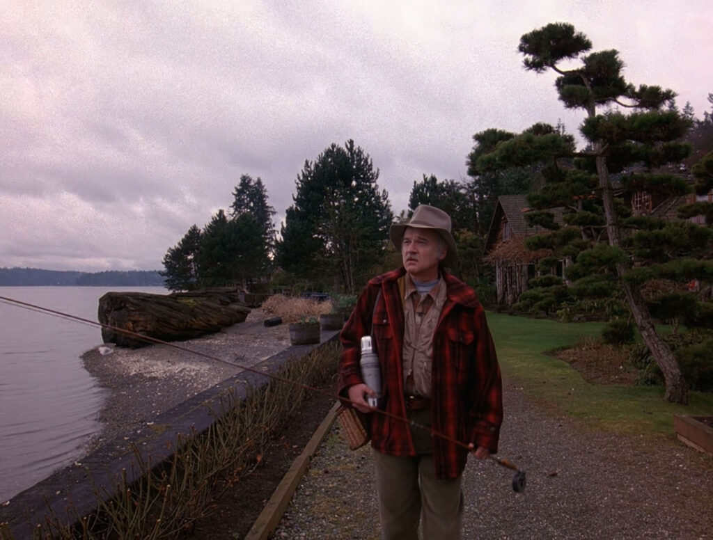 Pete Martell holding fishing gear on a path outside Blue Pine Lodge. The body of Laura Palmer wrapped in plastic is found near a giant log in the distance.