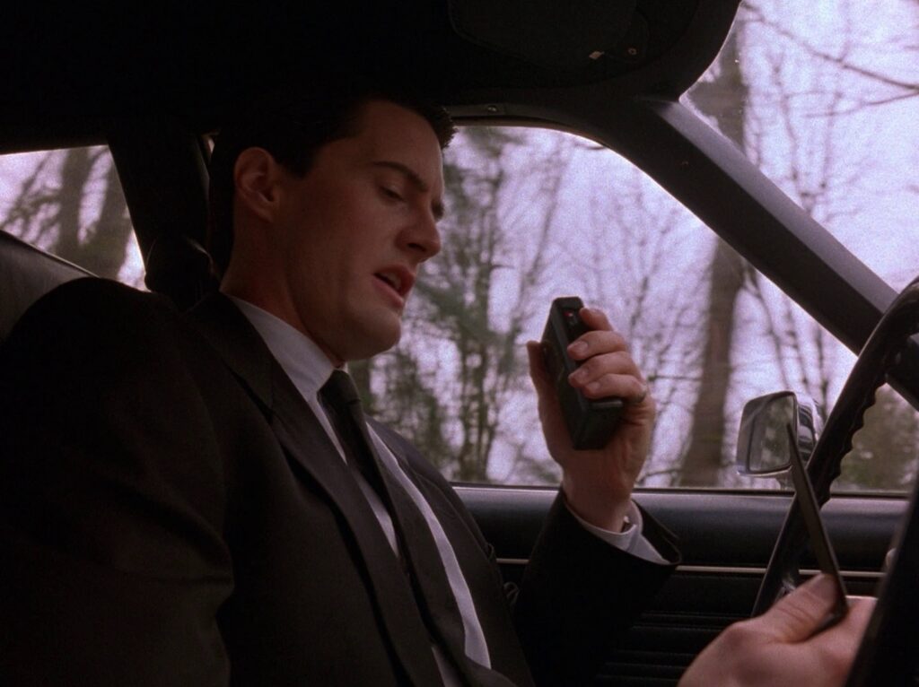 Agent Cooper driving a car while holding a tape recorder and checking his Dial Master