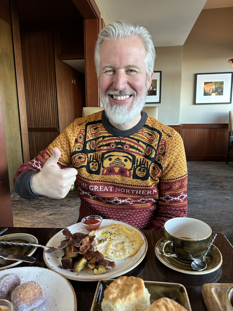 Steven showing a thumbs up at a breakfast table wearing a Great Northern Hotel sweater