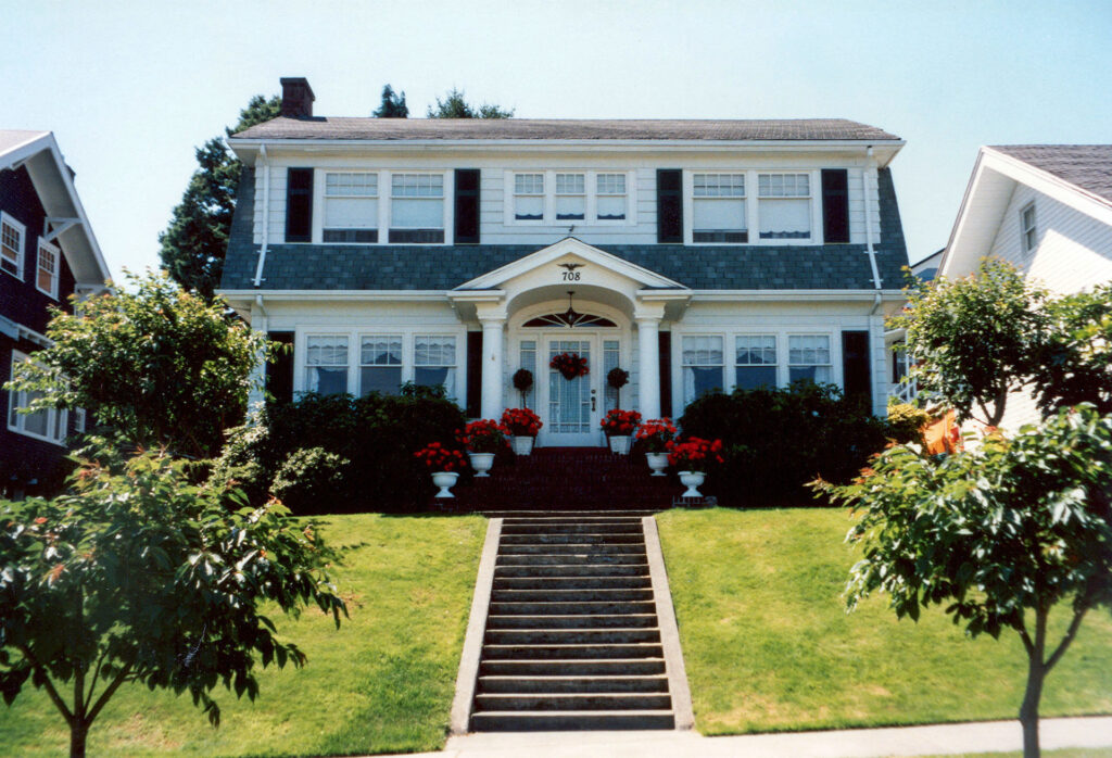 White house with bushes and small trees sitting on a small hill with concrete steps leading to the front door.