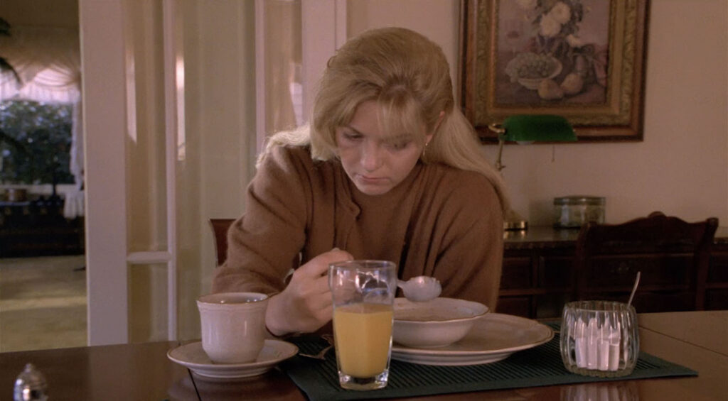 Laura Palmer sitting at a breakfast table holding a silver spoon above a bowl of soggy cereal. A glass of orange juice and coffee are on the table along with a bowl of sugar.