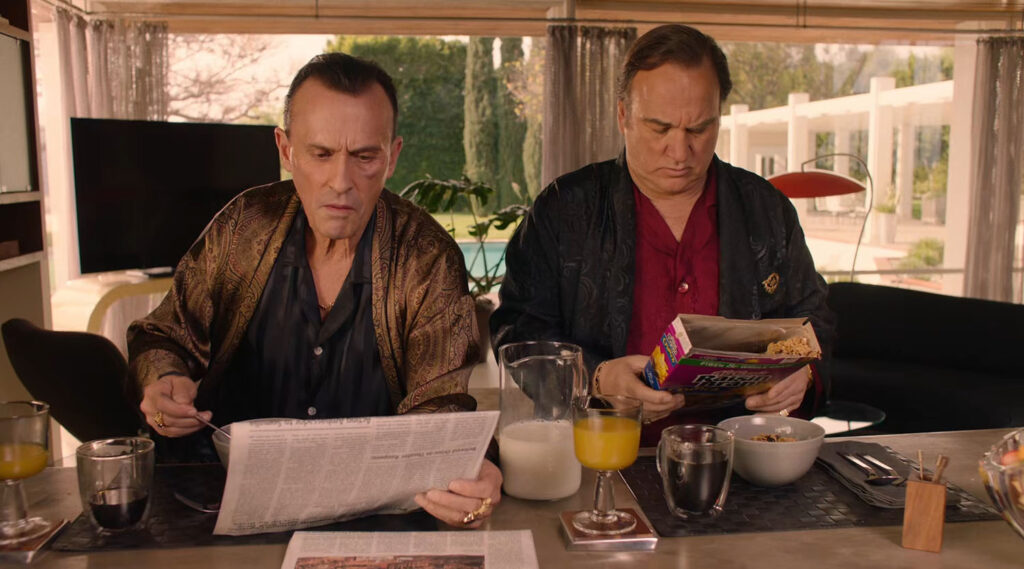 Two adult males eating breakfast at a counter wearing silk robes. One is holding a newspaper while the other is reading the back of a cereal box.
