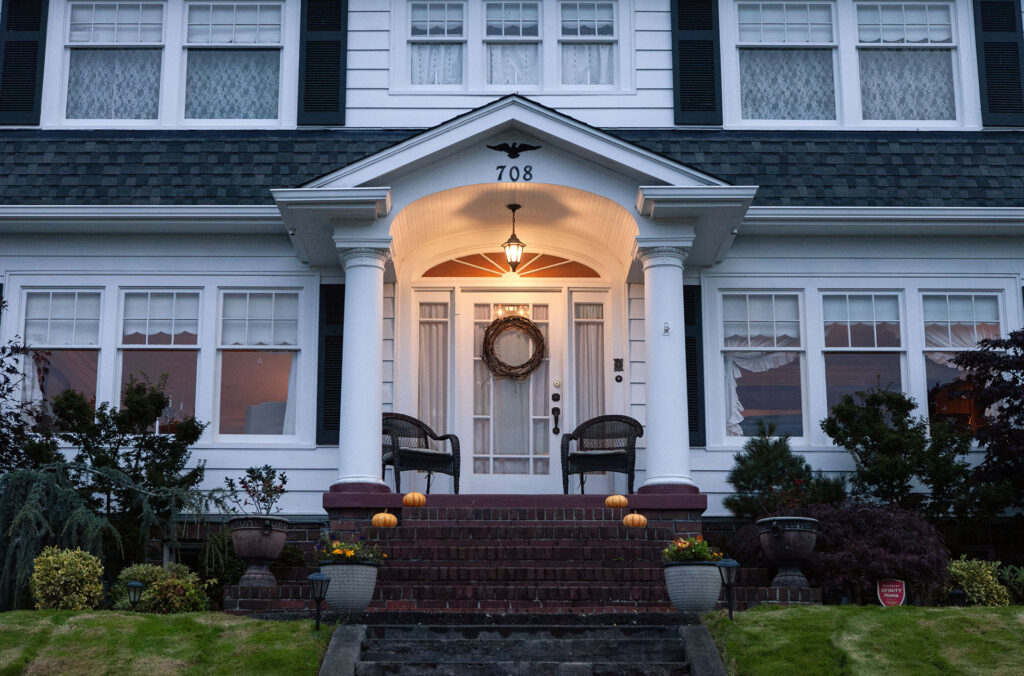White house on a hill with a porch light, two chairs on the front porch and a circular wreath hanging on the front door.