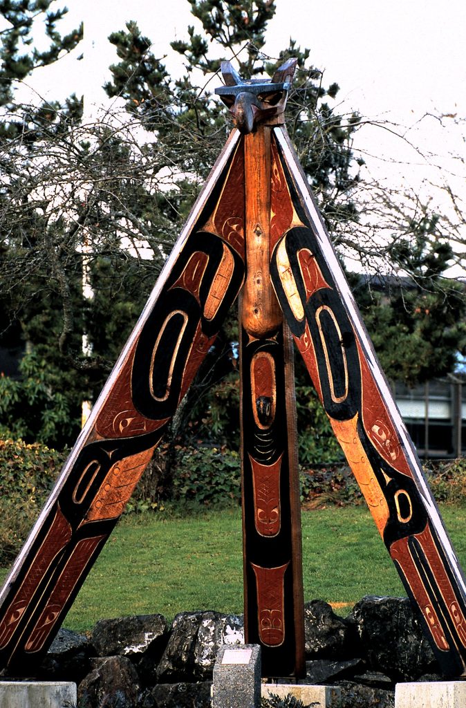 Wooden sculpture with three long carved poles that form a triangle.