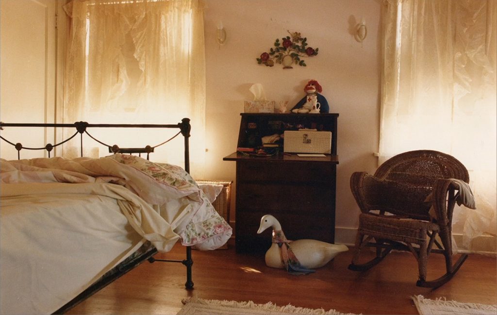 Laura Palmer's bedroom with unmade bed, small desk, a chair and two windows with curtains