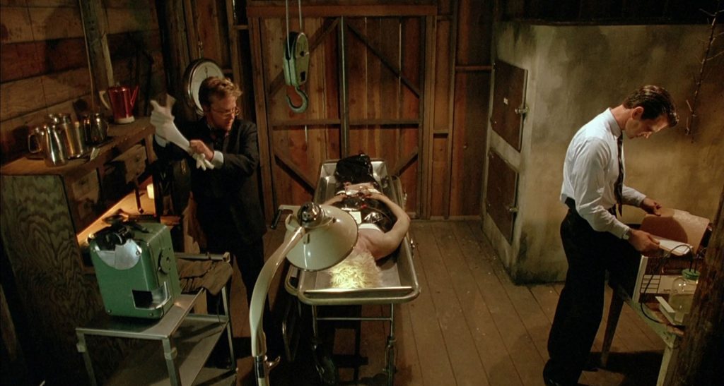 Interior of wooden shack with Agent Sam Stanley putting on rubber gloves while Agent Desmond looks through a file box. In the middle of the room is Teresa Banks' body on a metal slab.