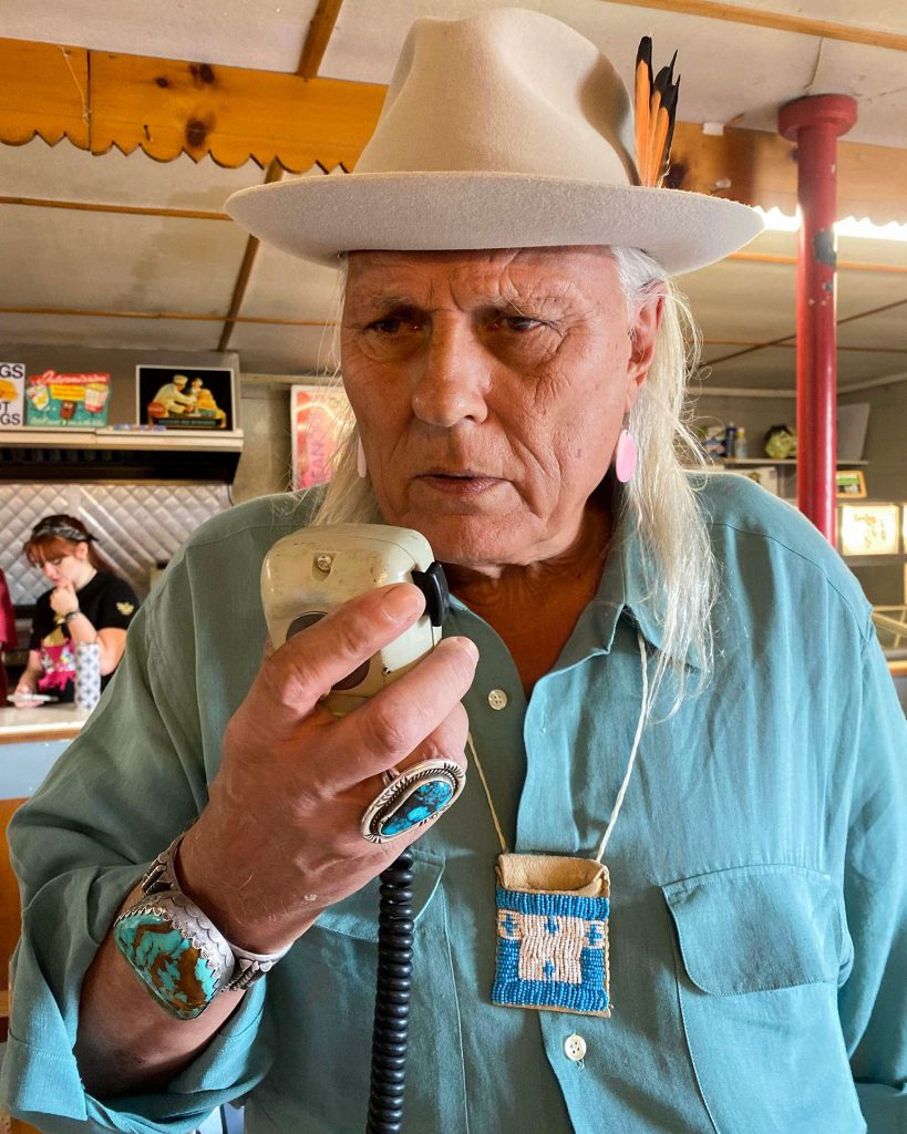 Michael Horse wearing a blue button down shirt and a hat holding a radio microphone