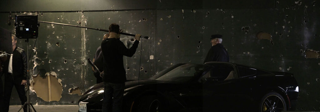 A Corvette Stingray with actors and a man holding a microphone boom against a wall full of holes.