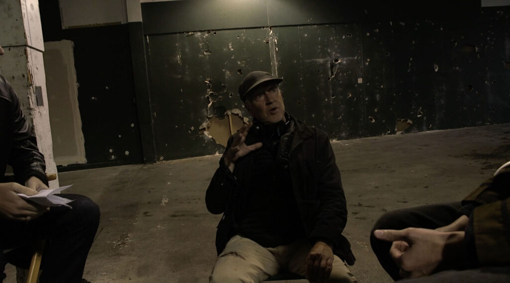 David Lynch seated in a garage with a hole filled wall behind him.
