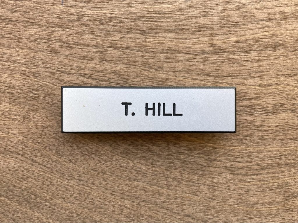 Silver name tag wit "T. Hill" printed in black lettering
