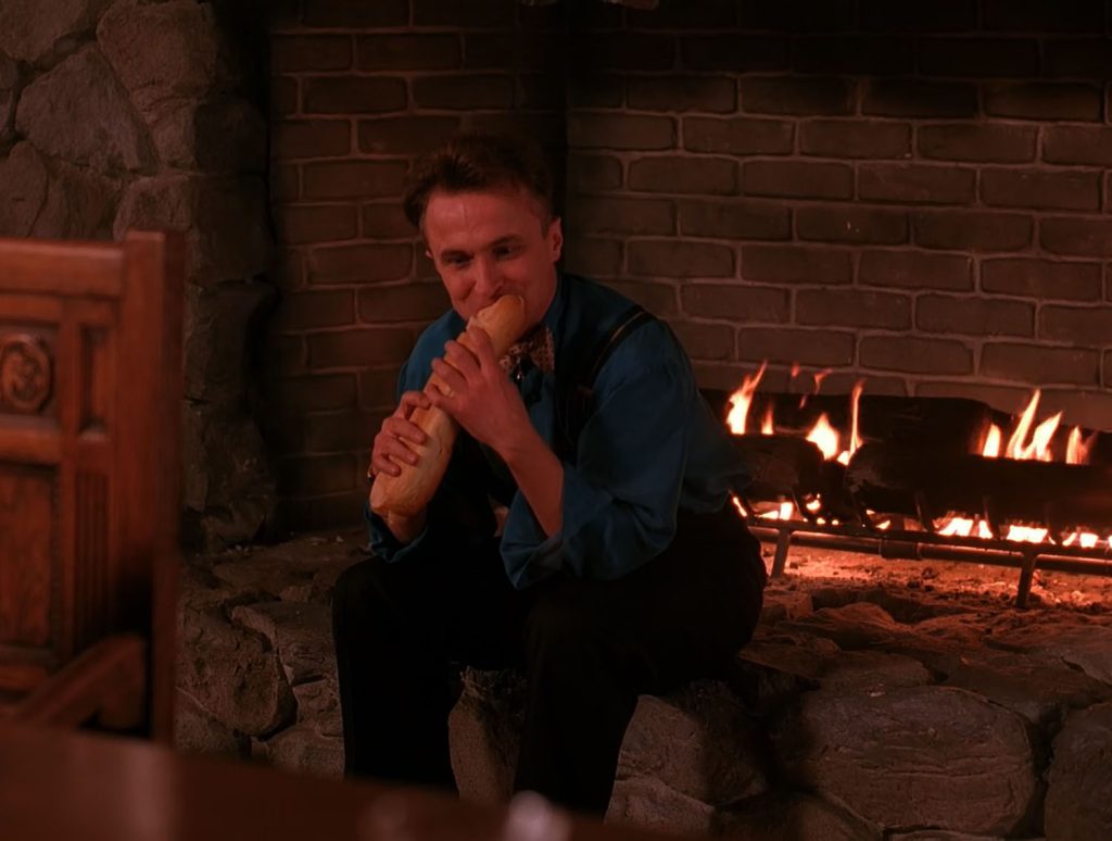 Jerry Horne sitting by fireplace holding a baguette