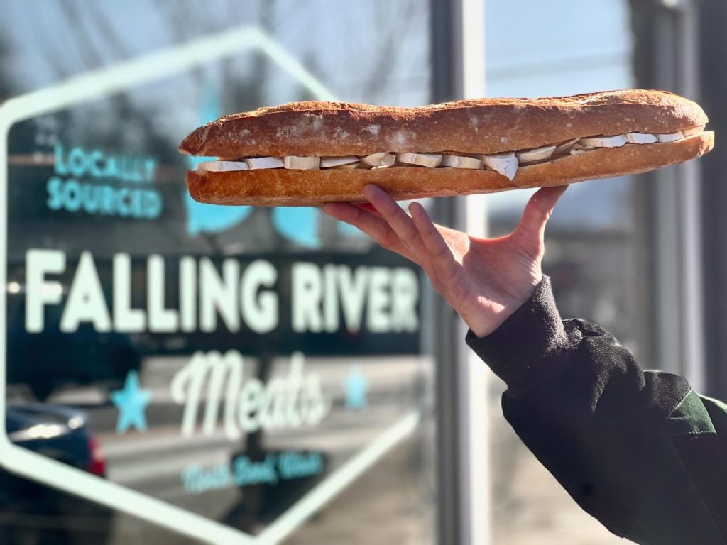 A person holding a baguette with Brie and butter in front of the Falling River Meats store in North Bend, WA