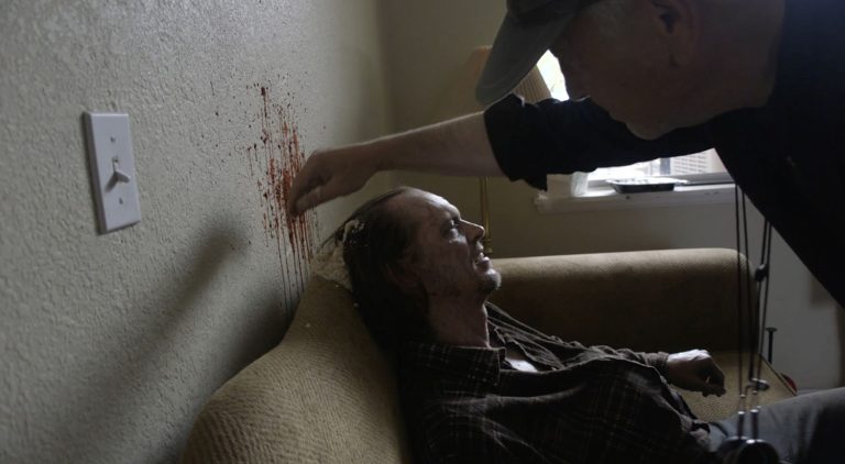 David Lynch applying make up to a Dead guy sitting in chair with a bullet wound to the head