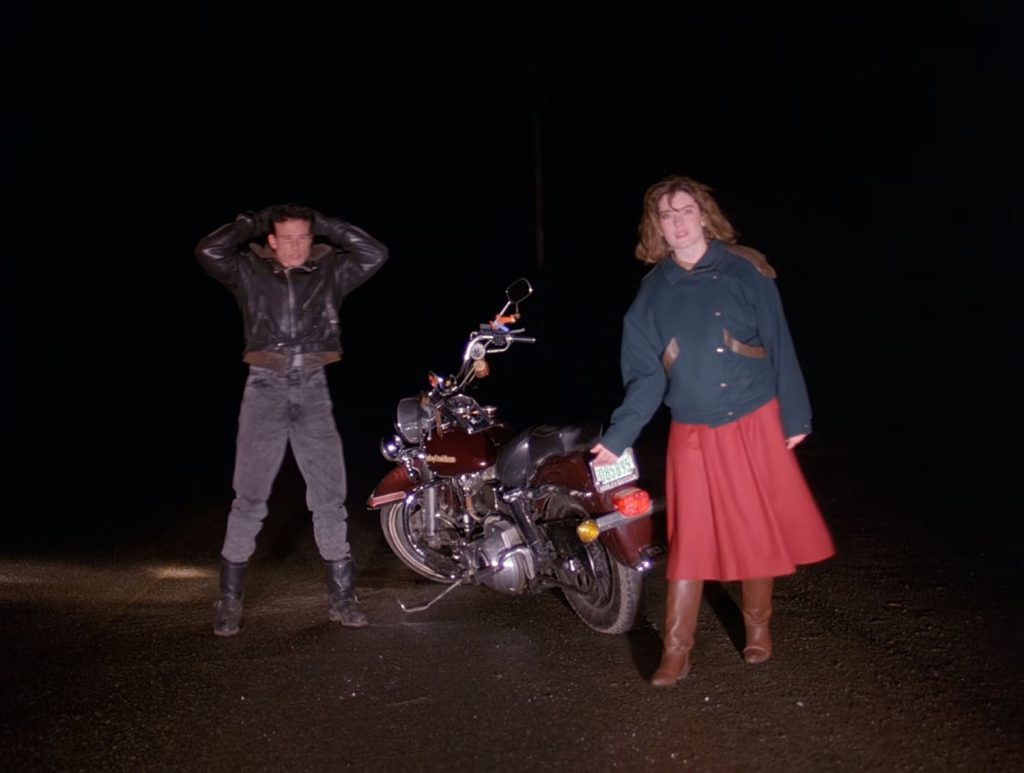 Donna Hayward standing on the road next to James Hurley's bike while James has his hands above his head