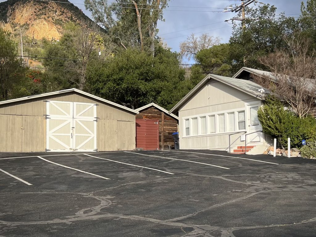 Two beige-colored barn structures located on a parking lot with tall trees in the distance