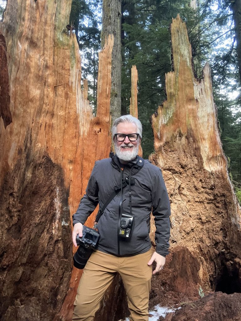 Steven with two cameras standing inside the remains of a fallen spruce tree in Olallie State Park