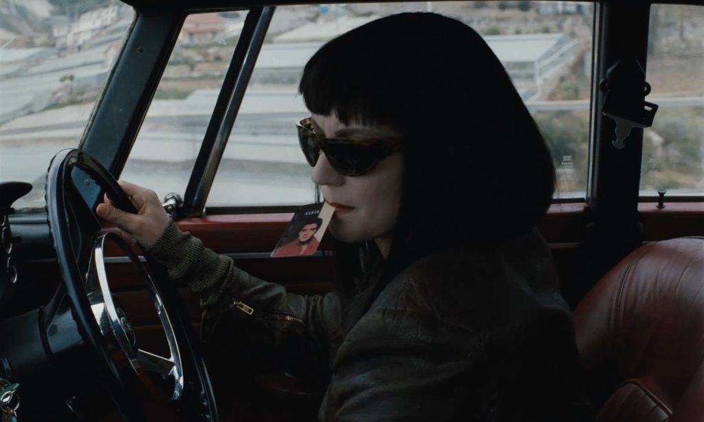 Clarie driving with Elvis card dangling from mouth