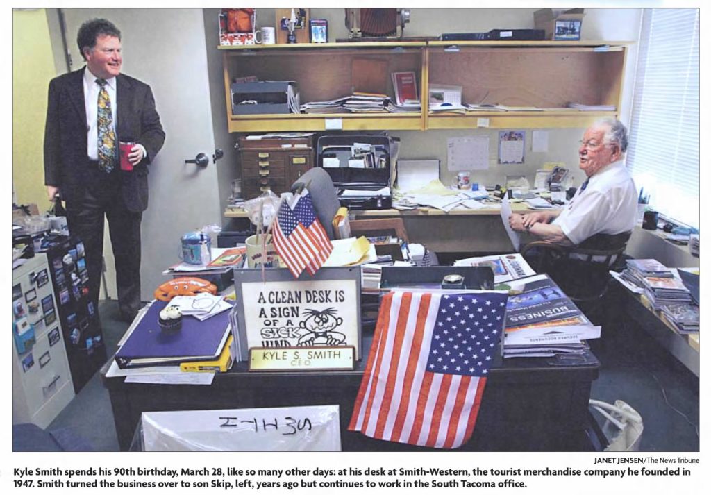 Kyle Smith sits at a desk talking with Skip Smith who is standing