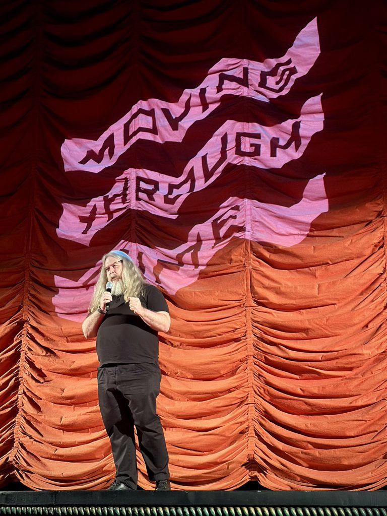 Daniel Knox on stage in front of a red curtain with the "Moving Through Time" logo